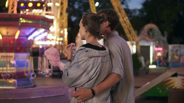 Backside View of a Young Dating Couple Walking By Funfair at Night Eating Cotton Candy alt