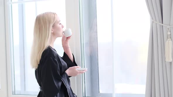 Young Beautiful Woman in Silk Black Robe Drinking Coffee Near the Window at Home alt