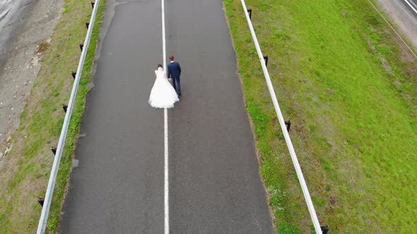 Newlyweds on Their Wedding Day Walk Along the Road alt