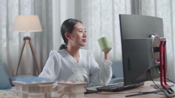 Asian Woman Engineer With The House Model Drinking Coffee While Working On A Desktop At Home alt