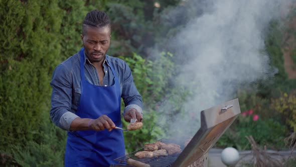 Confident Concentrated African American Man Taking Off Grilled Burger Patties From Barbecue Grid alt