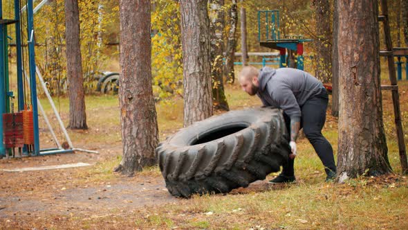 A Big Man Bodybuilder Turning Over the Tire on the Floor and Moving It - Training Outdoors alt