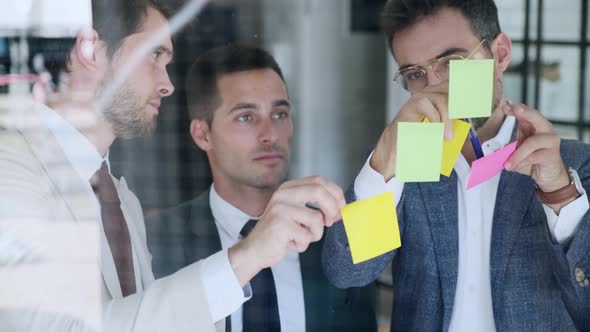 Businessmen brainstorming with post-its on glass pane alt