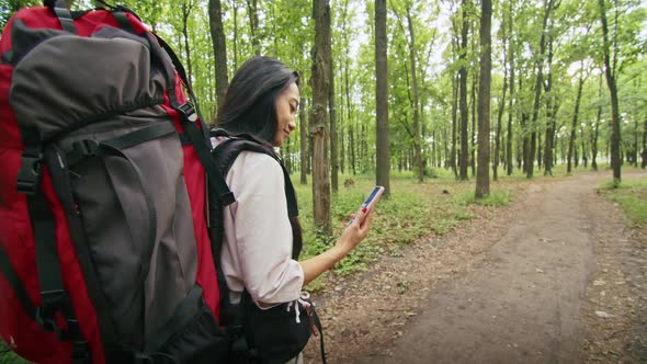 Happy Asian Woman with a Backpack Looking for a Way in the Forest Using a Phone Talking on a Video alt