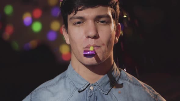Portrait of Happy Smiling Man in Birthday Hat Looking in the Camera with Noisemaker alt