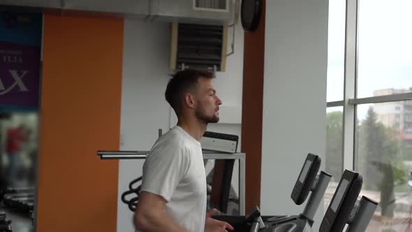 Slow Motion of a Young Man Running on Treadmill in Fitness Center. Side View.  alt