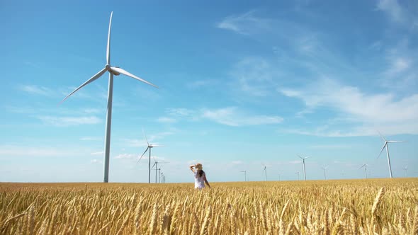 Beautiful girl walking on yellow field of wheat with windmills for electric power production alt