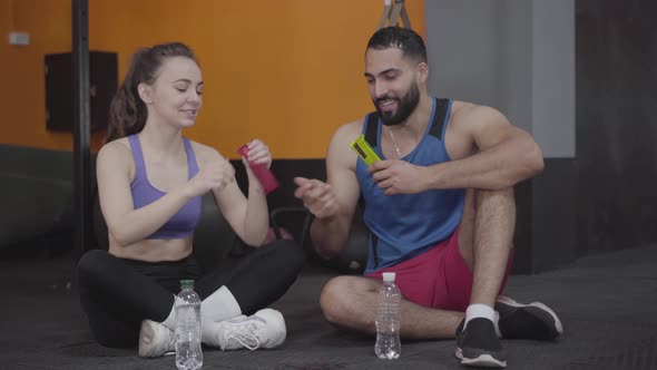 Positive Young Man and Woman Eating Protein Bars As Sitting in Gym and Talking alt