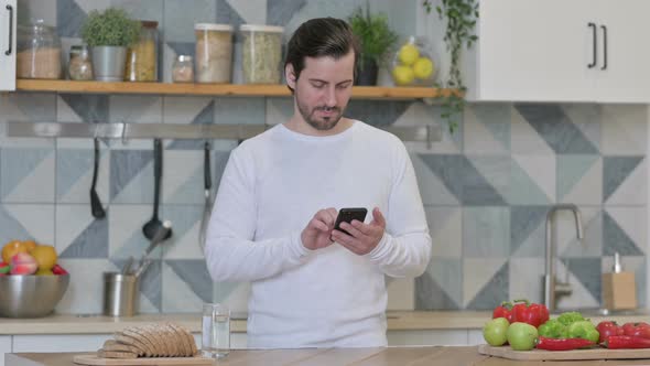 Young Man Using Smartphone While Standing in Kitchen alt
