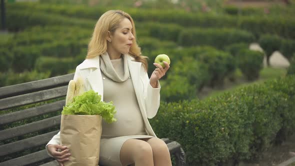 Smiling Expecting Lady Eating Fresh Apple on Bench With Grocery Bag, Health Care alt