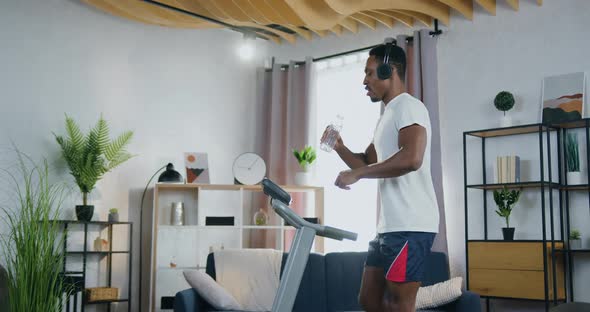 Man Listening Music in Headphones and Drink Water During Training on Treadmill at Home alt