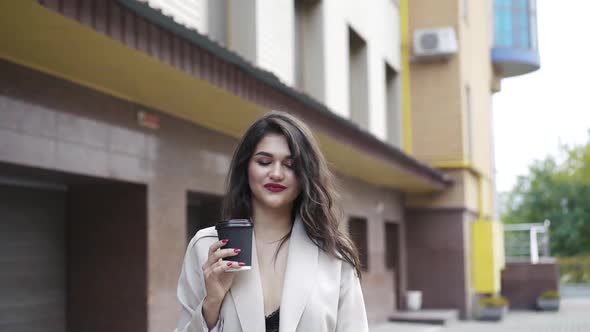 Brunette Dressed in Stylish Clothes Drinks a Beverage on the Street and Smiles alt