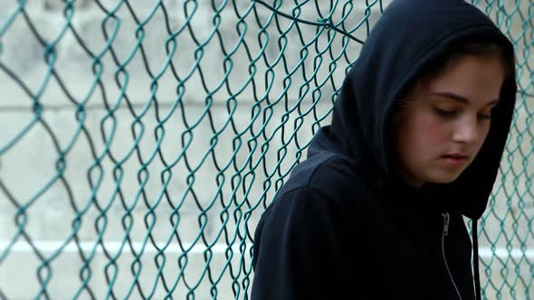 Sad schoolgirl in hooded leaning against fence alt