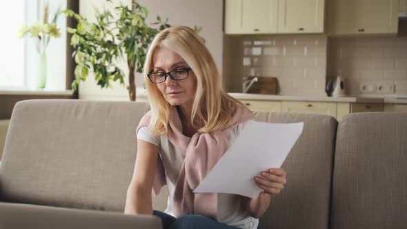 Mature Woman Sitting at Her Desk with Paper Letter Document and Laptop alt