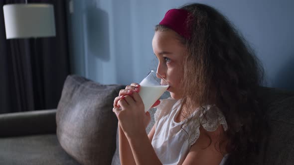 Positive Child Little Girl Drinks Milk While Sitting on Sofa at Home While Watching TV alt