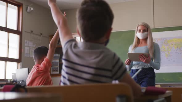 Female teacher wearing face mask pointing towards a boy raising his hands in class at school alt