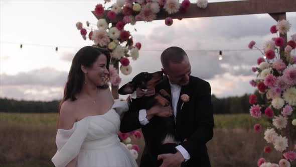 Funny Portrait of Laughing Newlyweds with a Dachshund in Their Arms Near a Flower Arch in the alt