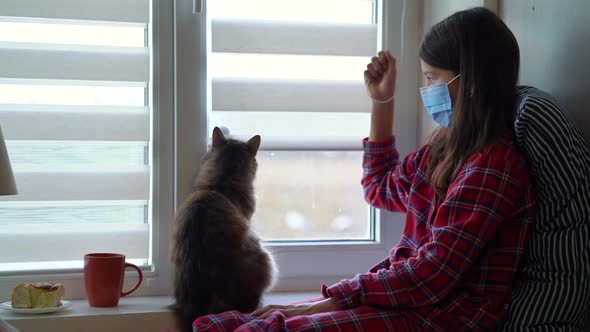 Girl in mask opening window blinds and cat watching alt