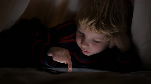 Young boy lying under blanket and watching cartoon on touch pad.