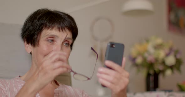 Happy Aged Woman Uses Phone Smiles Laughs Waves Her Hand and Sends Air Kiss alt