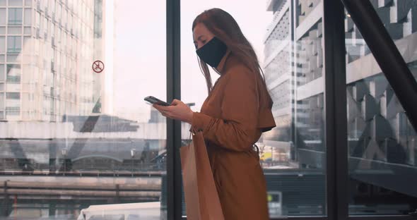 Side View Young Beautiful Business Woman in Face Mask Walks Using Mobile Office App at Mall Window alt
