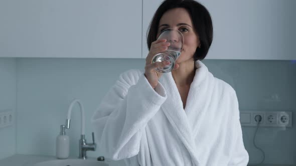 Thirsty smiling 35s woman in white bathrobe drinking water