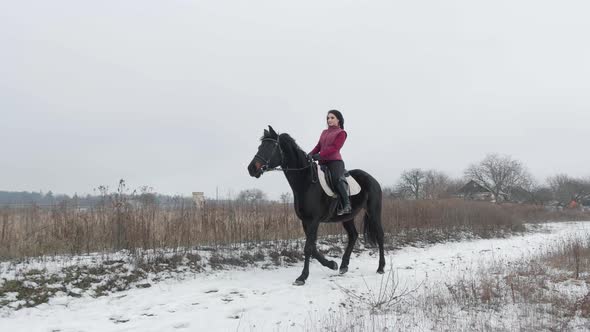 Young Brunette Woman Rides a Beautiful Black Horse on a Field or Snowcovered Farm in Winter alt