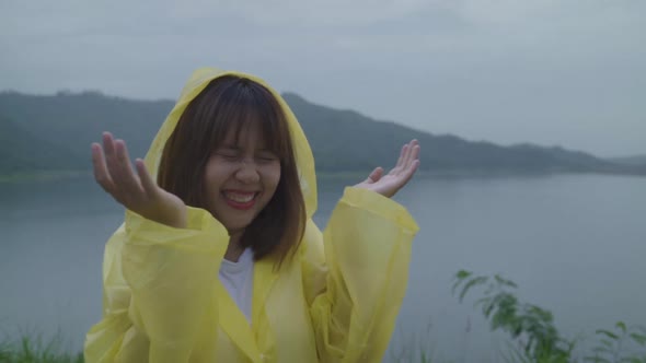 Young Asian woman feeling happy playing rain while wearing raincoat standing near lake. alt