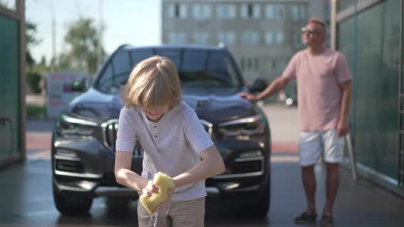 Cheerful Cute Boy Squeezing Water Off Sponge at Car Wash Service Standing at Clean Automobile with alt
