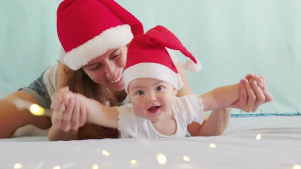 Smiling Santa Child Lying in Santa Claus Costume, in Red Hat with Mother. alt