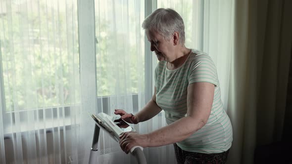 Old Woman Walks on a Treadmill in a Front of the Window alt