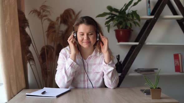 Young Beautiful Caucasian Woman Sits at Home at a Desk Listens to Music on Headphones and Dances alt