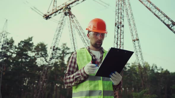Electrical Equipment Worker Near High Voltage Tower Using Walkie Talkie and Clipboard alt