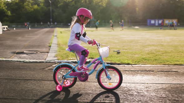 Cute Light Hair Little Girl in Pink Helmet in Elbow and Knee Pads Rides a Bicycle at the Stadium