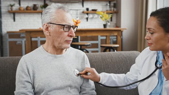 Woman Doctor Examining Aged Man with Help of Phonendoscope Listening to His Lungs Sitting on Sofa alt