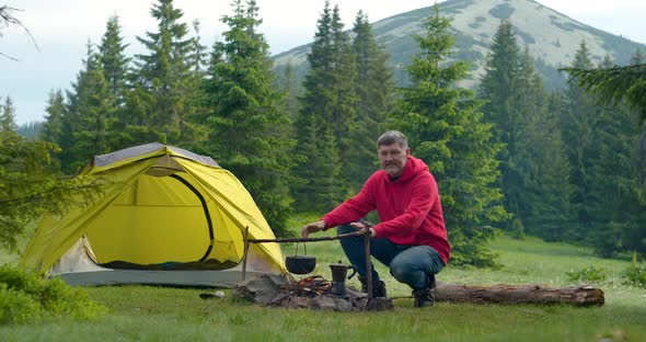 Bearded Male Tourist Warms His Hands Over a Bonfire in the Mountains alt