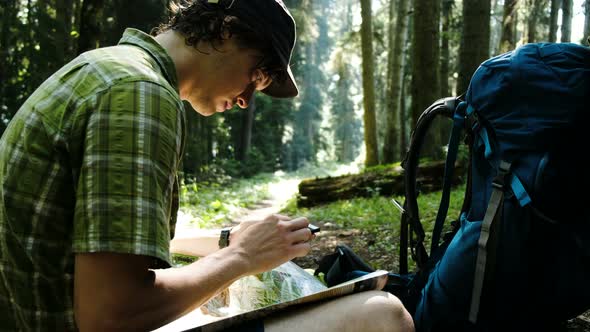 Tourist with a Backpack Sits in the Forest and Uses an Electronic Map on His Phone, Checks the Route alt