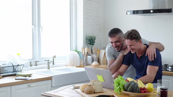 Happy gay male couple browsing internet together in kitchen at home
