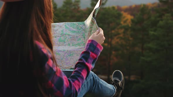 A Young Woman is Sitting on a Rock and Holding a Map in Her Hands alt