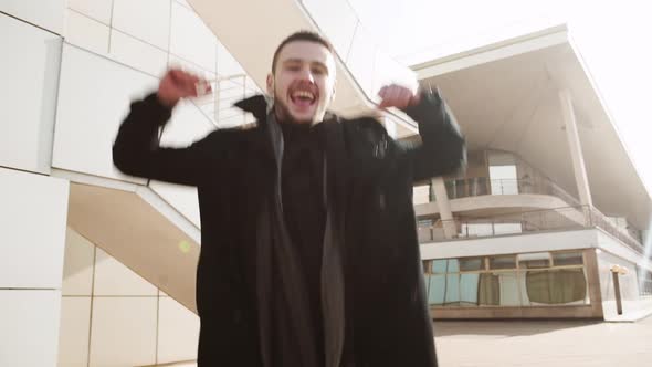 Young Handsome Caucasian Male in Black Coat Jumping for Joy and Smiling Outdoors alt