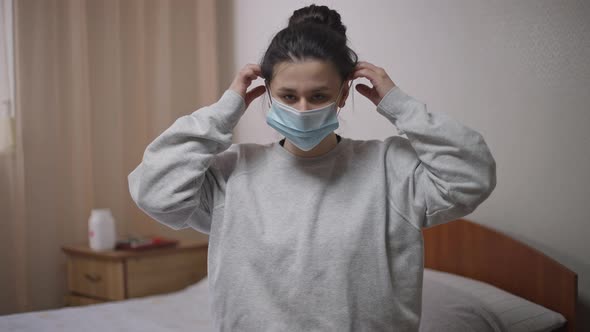 Young Woman Putting on Coronavirus Face Mask Looking at Camera Sitting on Bed Indoors alt