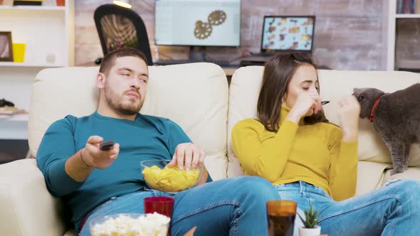 Bearded Man Eating Chips and Using Remote Control To Change Tv Channels alt