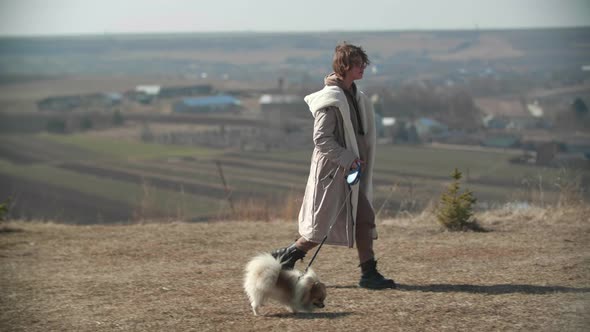 A Young Teenager Girl is Walking with a Small White Pomeranian Spitz on the Hill alt