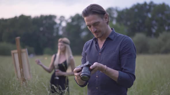 Happy Handsome Photographer Looking Through Photos on Camera As Blurred Beautiful Artist Painting at alt