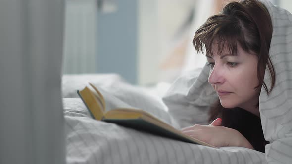 Woman Lies on the Bed and Reads a Book Covered with a Blanket