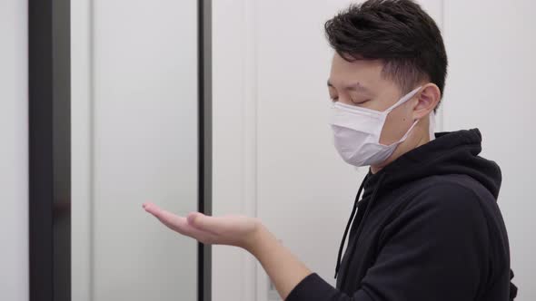A Young Asian Man in a Face Mask Applies Disinfection Gel on His Hands and Looks in a Mirror alt