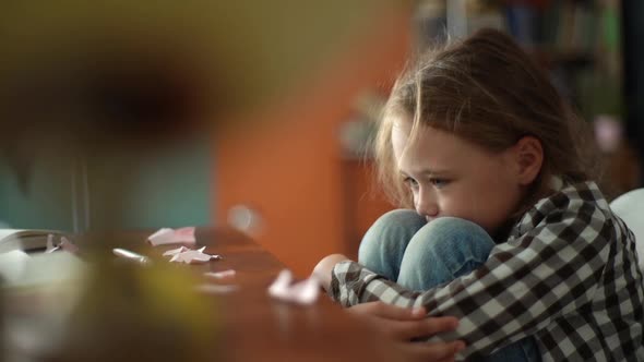 Side View of Exhausted Upset Primary Child School Girl Sitting Alone Hugging Knees in Front of Desk