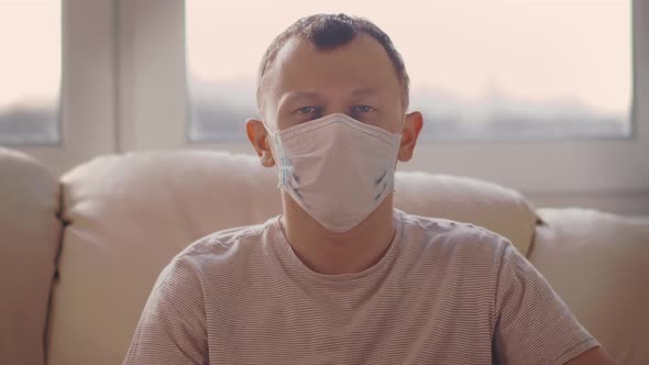 A Man in a Medical Mask Sits at Home Alone in Isolation, Looking at the Camera alt
