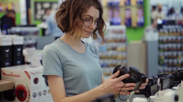 Young Woman in Glasses Shopping for a New Photocamera in the Electronics Store alt
