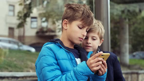 Two Boys Are Sitting At The Bus Stop. Playing Smartphones. alt
